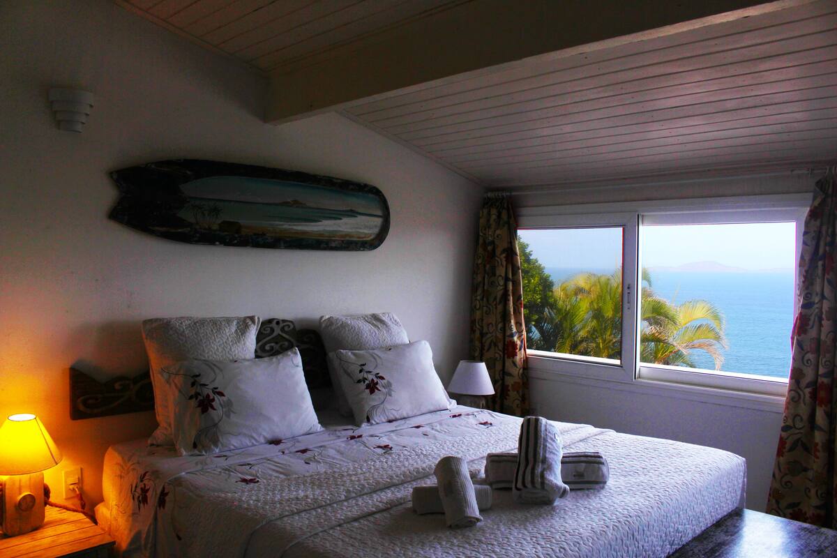Une chambre à coucher avec un lit, une table de chevet, une lampe, un tableau et une fenêtre donnant sur un beau paysage.