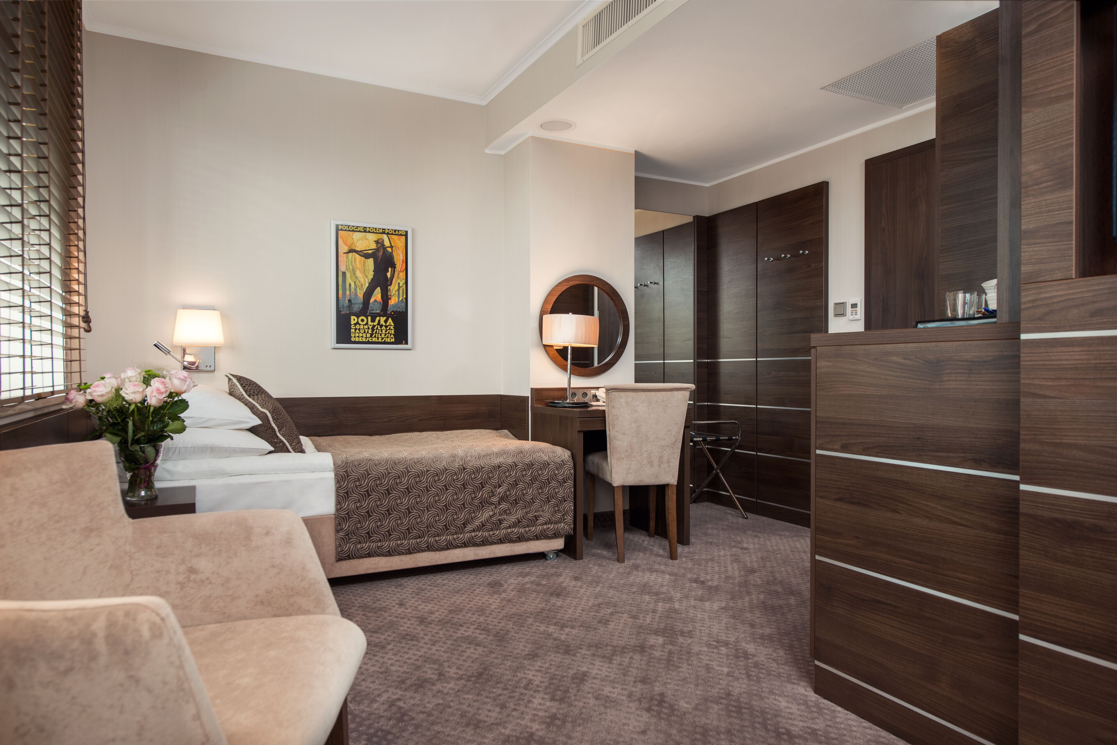 A hotel room with a bed, a desk, a chair, a lamp, a poster on the wall, and a large wardrobe.