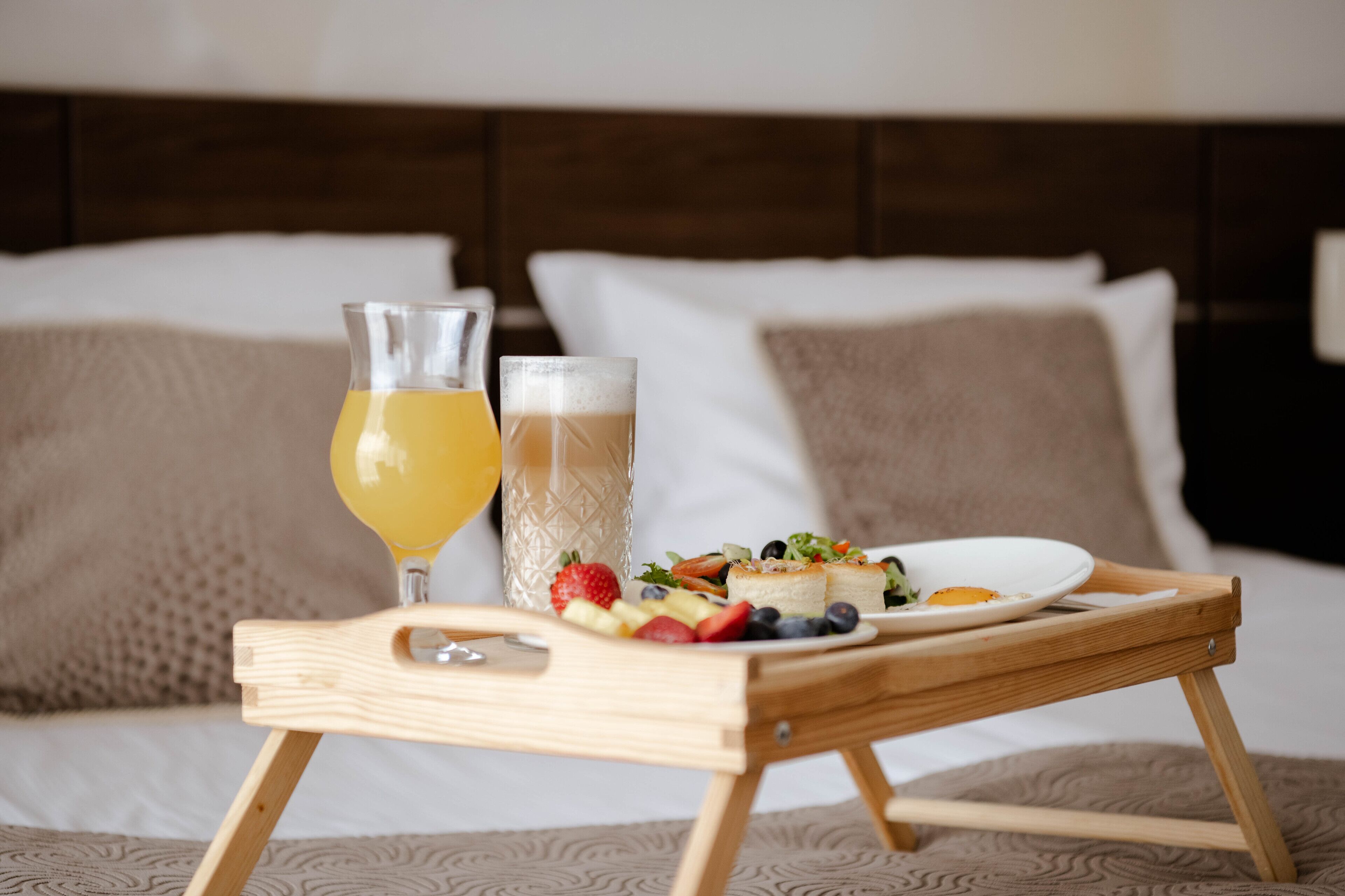 A wooden tray with a glass of orange juice, a glass of beer, and a plate of food on a bed with pillows.