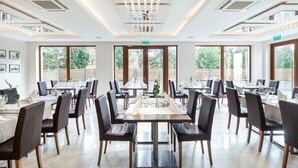 A spacious dining area with multiple tables and chairs, large windows with a view of greenery, and a modern chandelier. - Hotel Różany Gaj - Destigo Hotels (Gdynia)