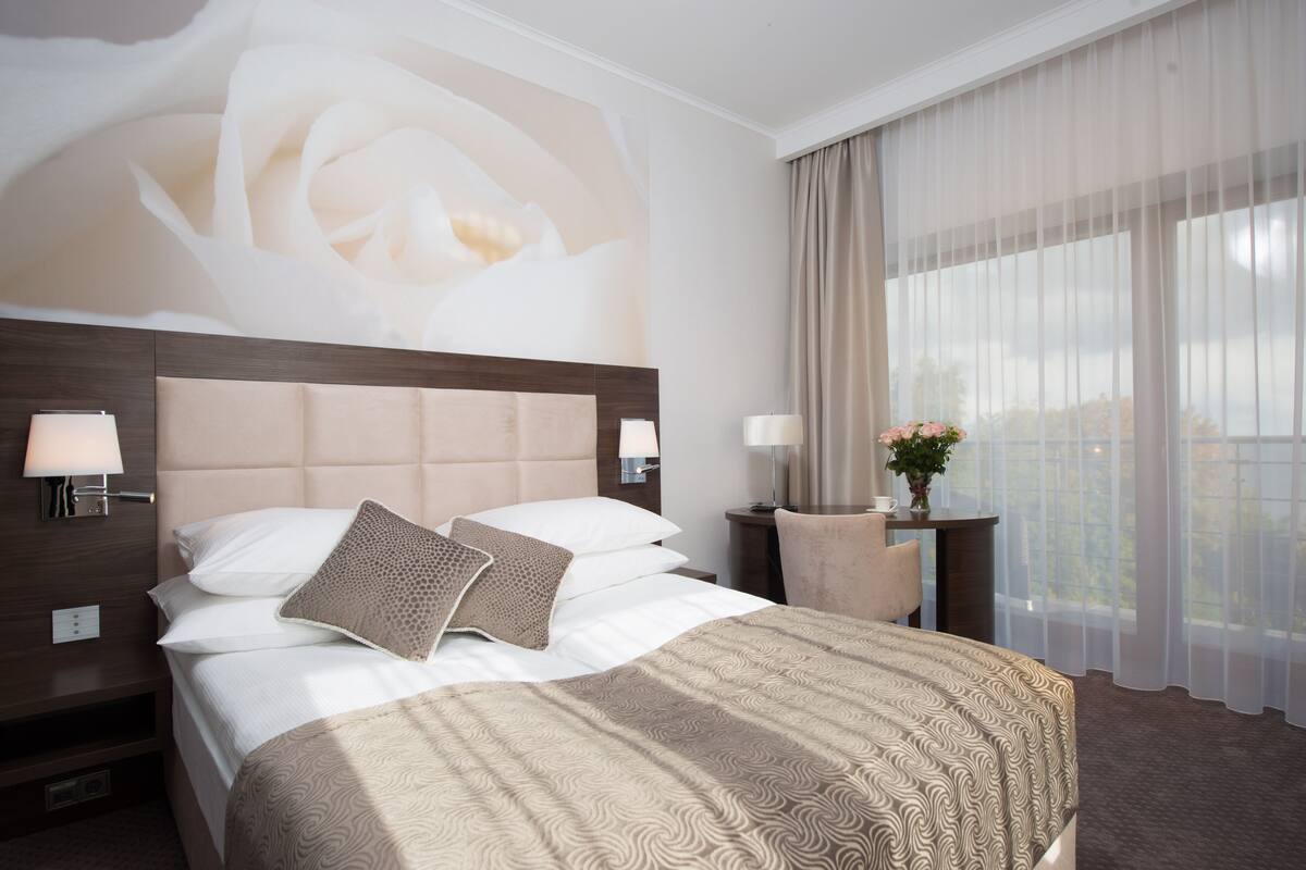 A hotel room with a large bed, a nightstand with lamps, a small table with a vase of flowers, and a view of the outdoors through sheer curtains.