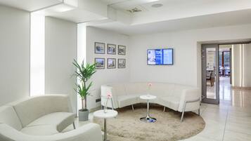 A modern, well-lit waiting area with a curved white sofa, a small round table with a potted plant, and a television mounted on the wall.
