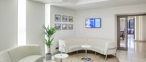 A modern, well-lit waiting area with a curved white sofa, a small round table with a potted plant, and a television mounted on the wall.