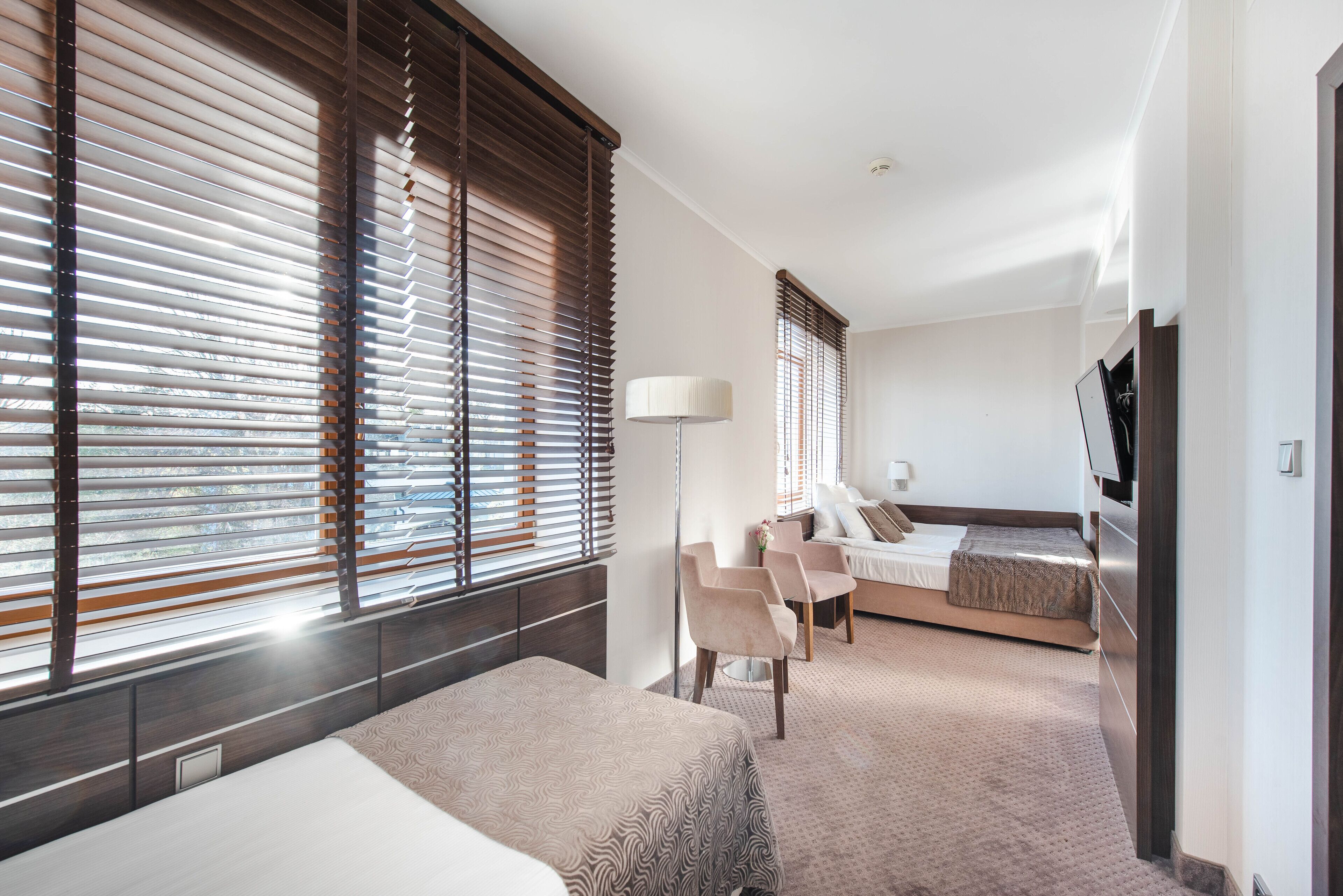 Ein Hotelzimmer mit einem Bett, einem Schreibtisch, einem Sessel, einem Fernseher und großen Fenstern mit Jalousien.