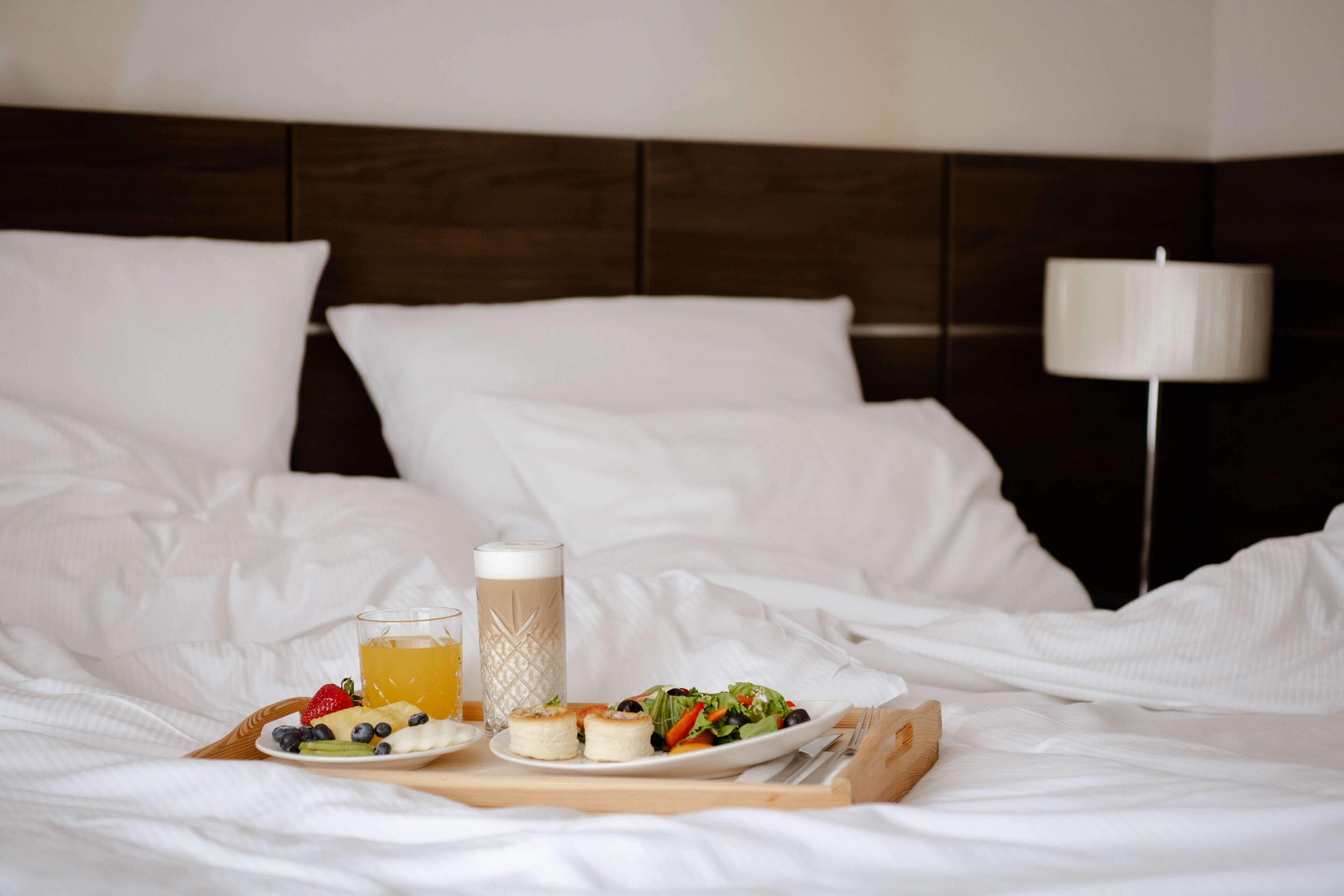 A neatly made bed with white linens, a wooden headboard, a bedside lamp, and a tray with breakfast including fruits, a drink, and a plate of food.