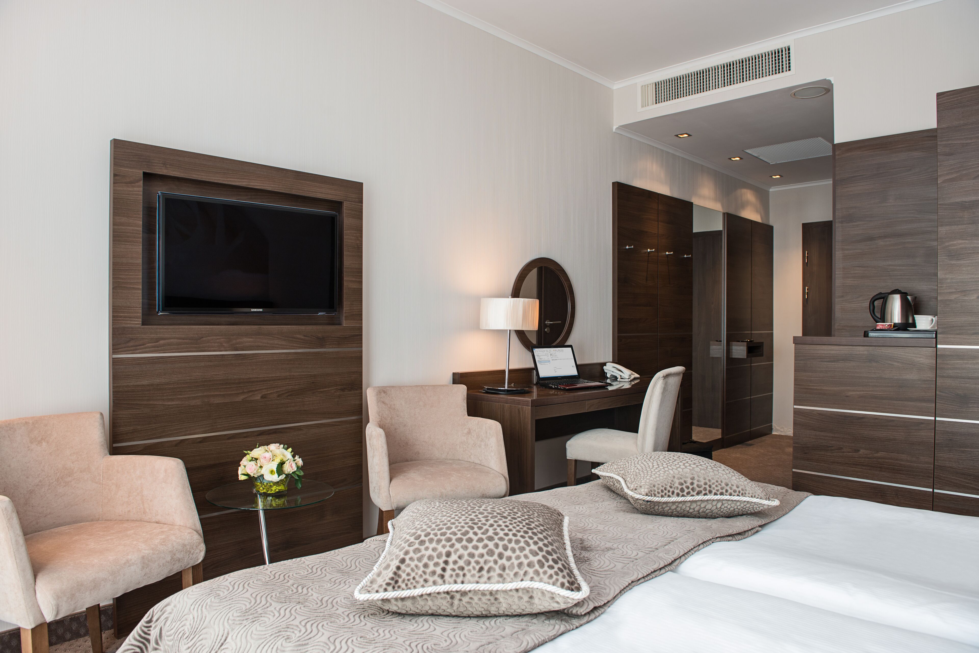 A modern hotel room with a flat-screen TV mounted on the wall, a desk with a lamp, two armchairs, a bed with decorative pillows, and a wardrobe.