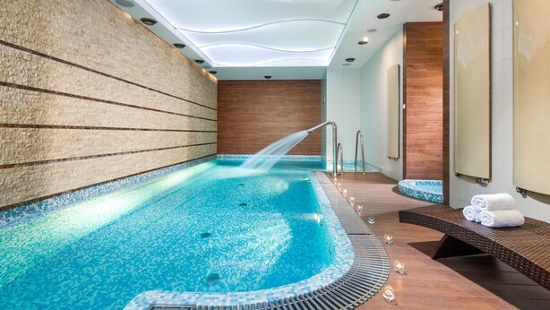 A modern indoor swimming pool with a clear water feature, a wooden wall, and a seating area with towels. - Hotel Różany Gaj - Destigo Hotels (Gdynia)