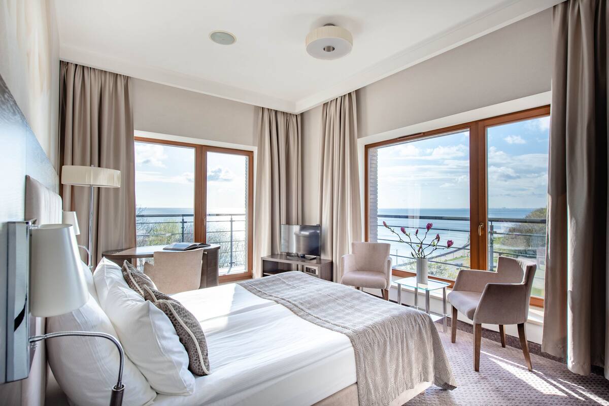 A hotel room with a large bed, a TV, a desk, and a balcony with a view of the sea.