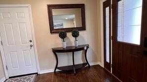 Um corredor com piso de madeira, uma porta branca, um console de madeira escura com duas pequenas plantas em vasos, um espelho grande acima da mesa e uma porta de madeira com janela à direita.