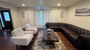 Uma sala de estar com sofá seccional, duas poltronas, uma mesa de centro e uma janela grande com cortinas.