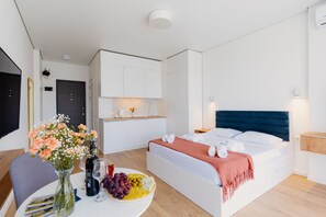 Una habitación de hotel moderna con una cama, una pequeña mesa de comedor con una bandeja de frutas, un florero con flores y vistas a la cocina.