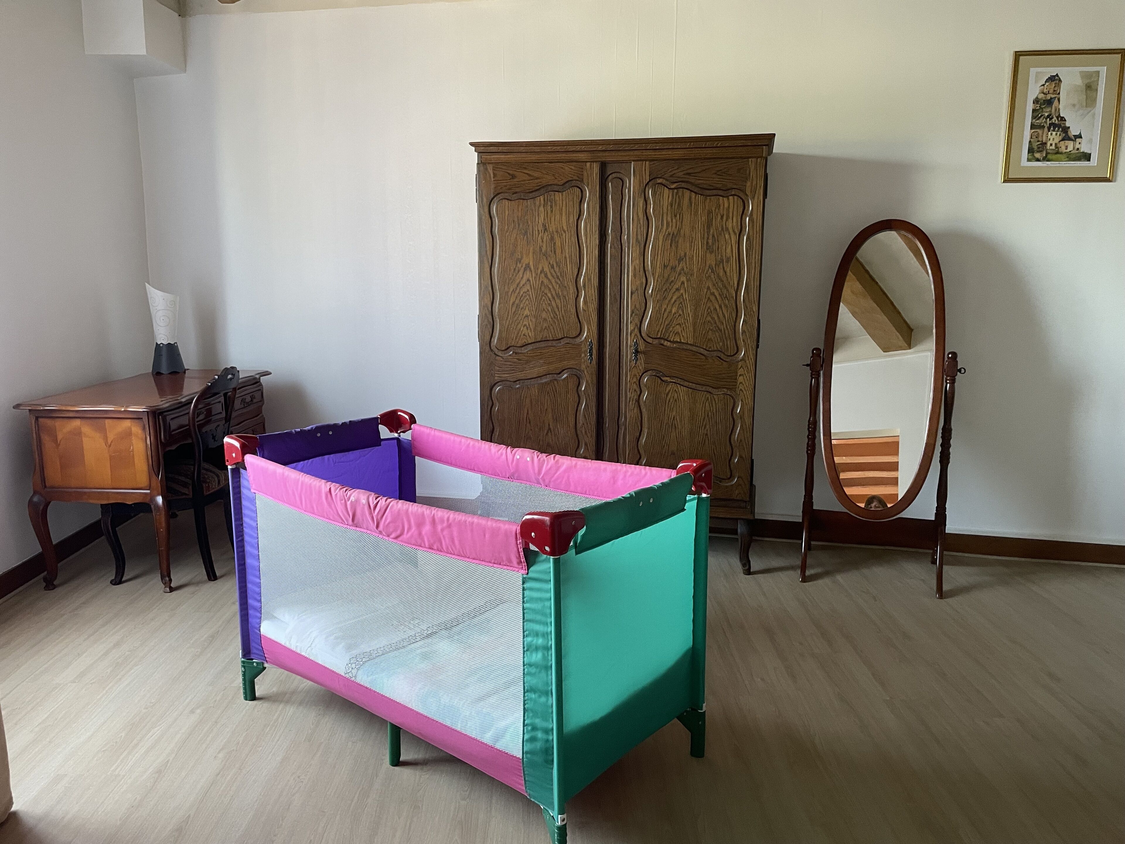 A room with a baby playpen, a wooden wardrobe, a mirror, and a desk with a lamp.