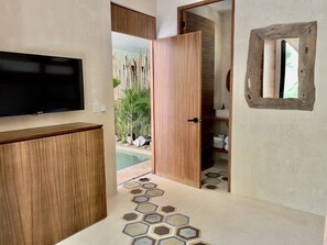 Ein Zimmer mit einem Holzschrank, einem an der Wand montierten Fernseher, einer Holztür, die ins Badezimmer führt, und einem hexagonförmig gemusterten Boden.