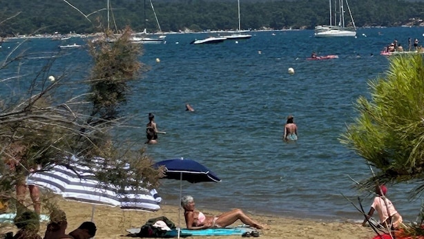 A beach with people sunbathing and swimming, boats in the water, and a mountainous backdrop.