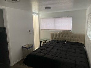 Un dormitorio con cama, una ventana con persianas, una mesita y un cubo de basura.