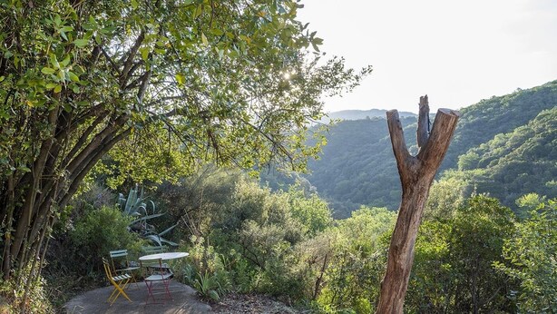 Un espace extérieur aménagé dans un style rustique, avec une table et des chaises, entouré d’une végétation luxuriante et offrant une vue sur une vallée boisée.