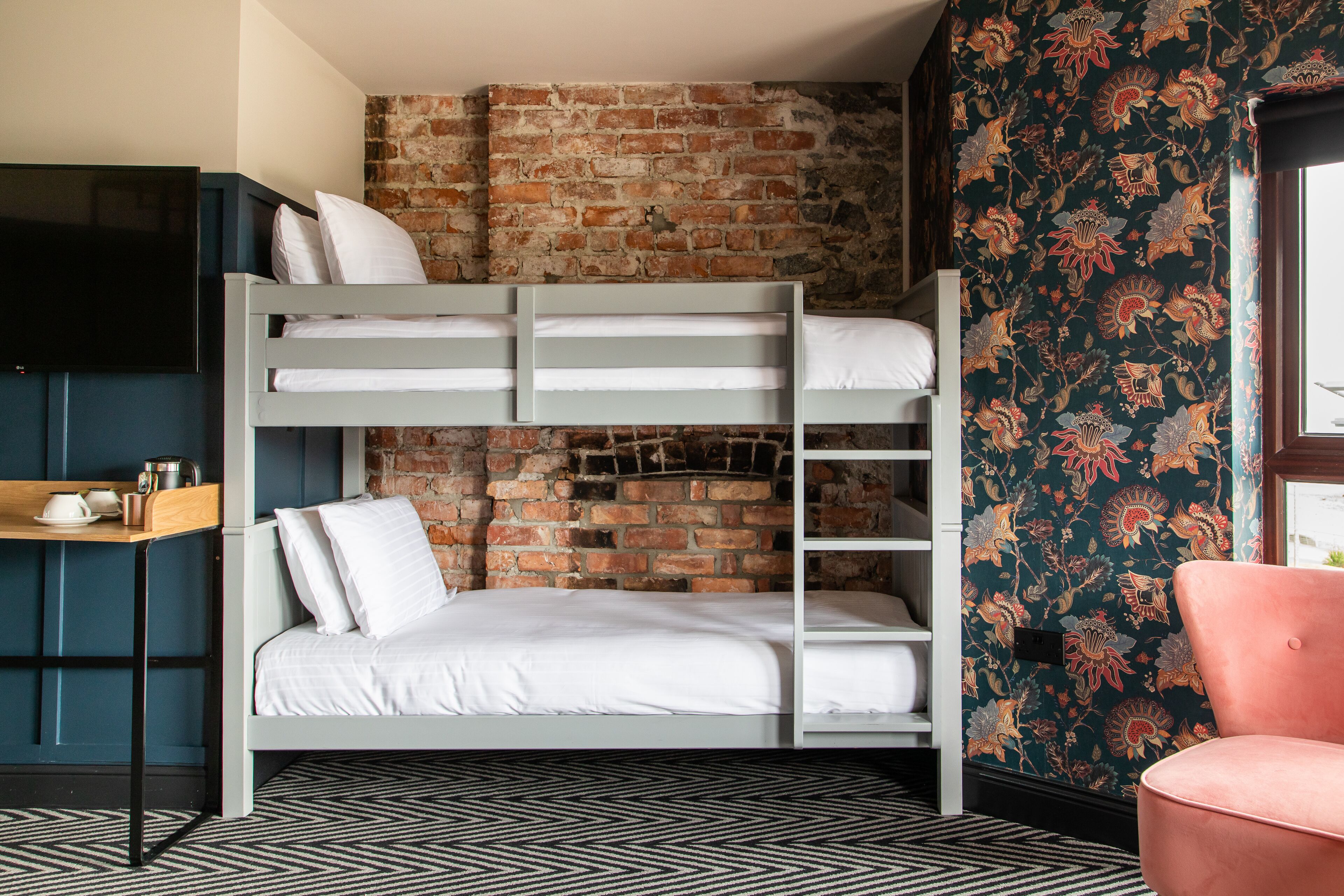 Una cama litera con ropa blanca, una pared de ladrillo, papel tapiz floral, una silla rosa y un televisor sobre un mueble.