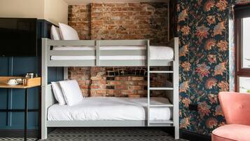 Una cama litera con ropa blanca, una pared de ladrillo, papel tapiz floral, una silla rosa y un televisor sobre un mueble.