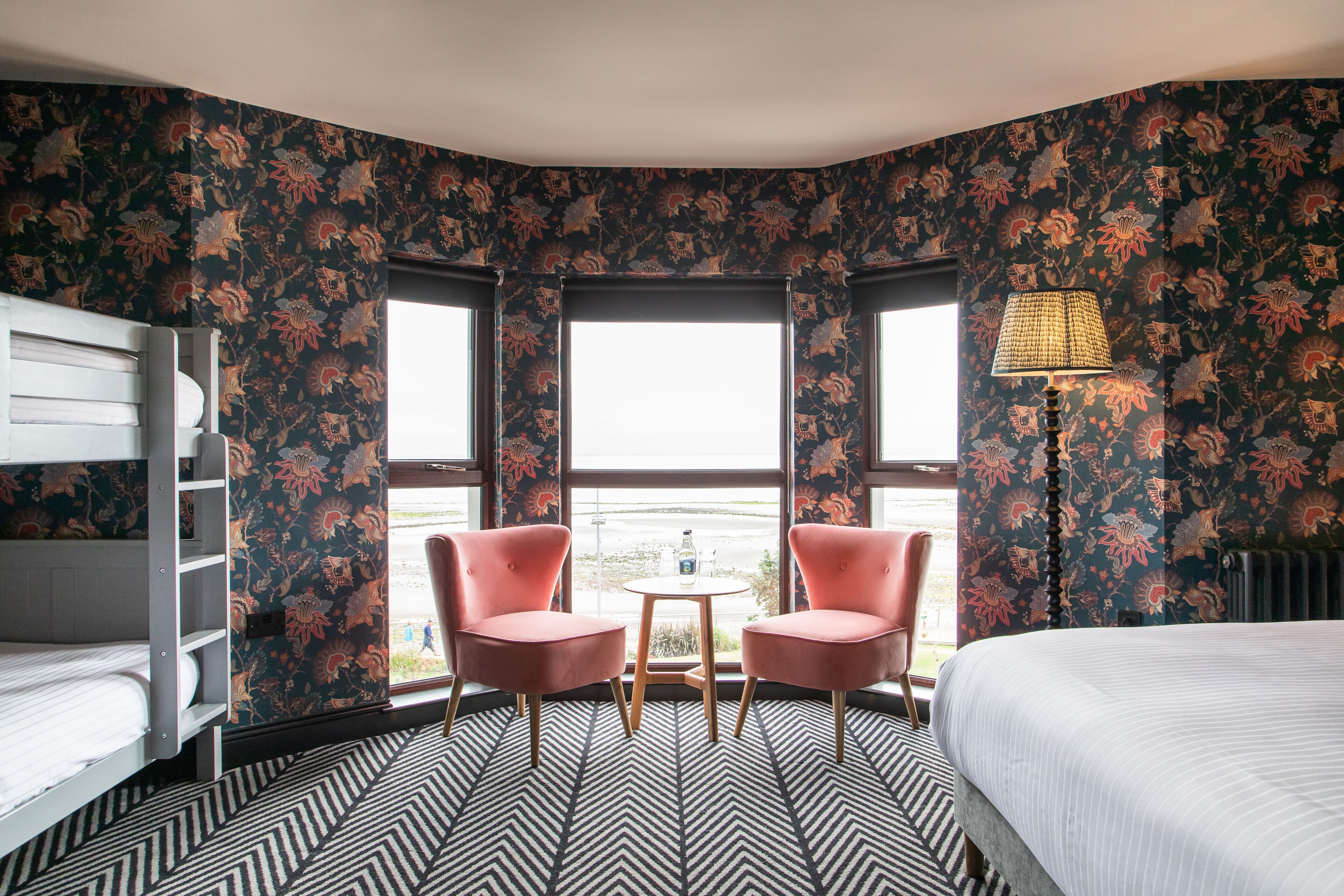 A room with a bunk bed, two pink chairs, a small table, and a lamp.