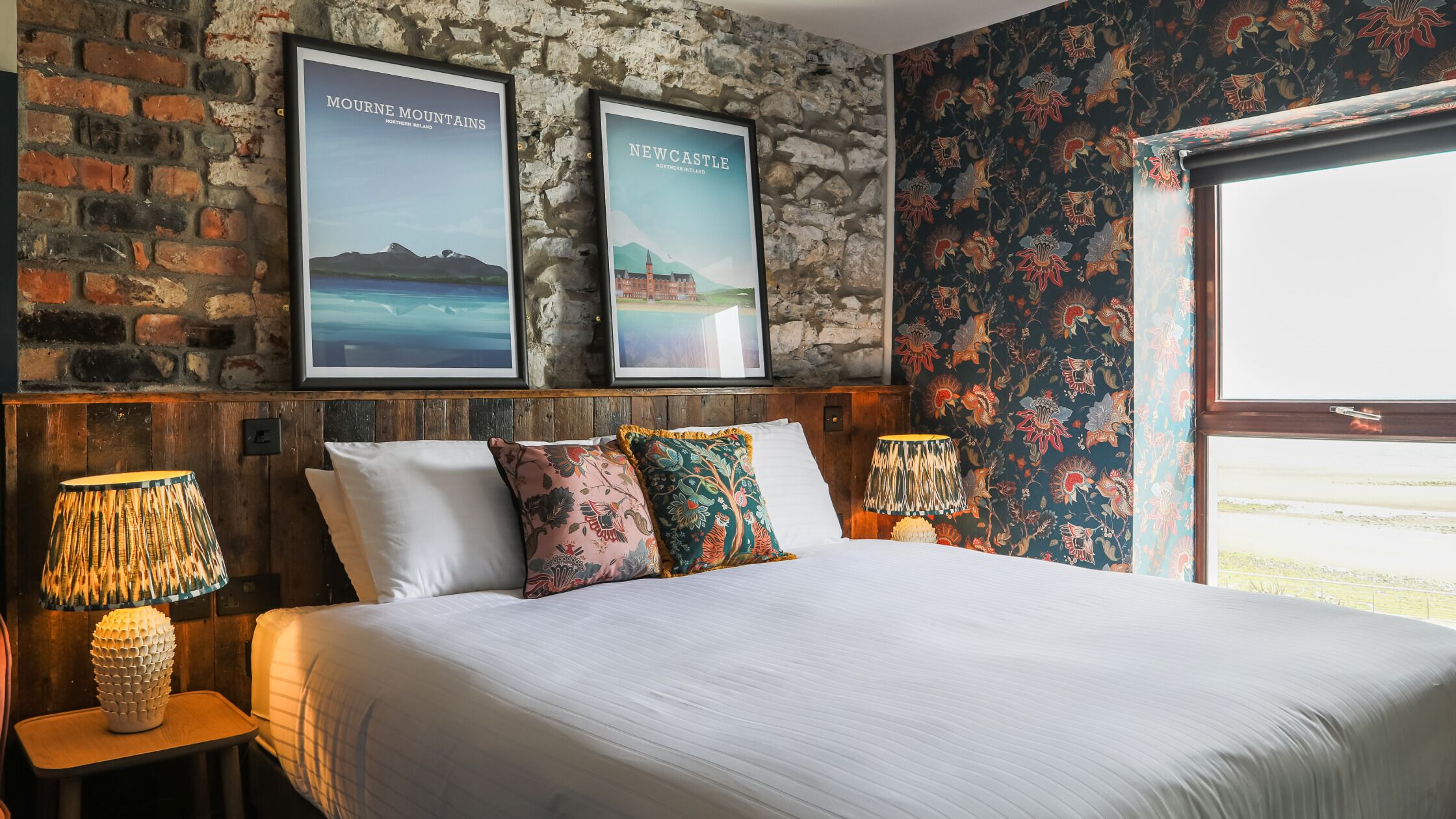 A bed with white linens, two framed posters of Northern Ireland landscapes, a floral wallpaper, and a window with a view.