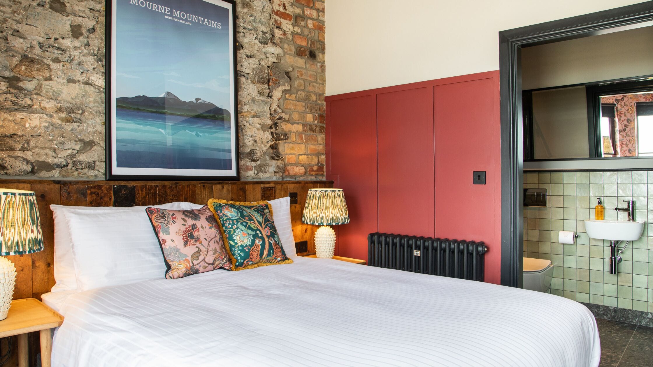 A hotel room with a large bed, a brick wall, a poster of the Mourne Mountains, a red cabinet, and a bathroom with a sink and mirror.