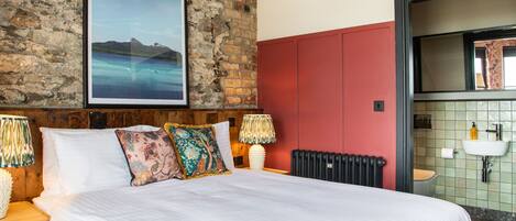 A hotel room with a large bed, a brick wall, a poster of the Mourne Mountains, a red cabinet, and a bathroom with a sink and mirror.
