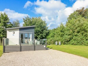A small, modern mobile home with a glass door and a deck, surrounded by a gravel driveway and lush greenery. - Number 15 (Carnforth)