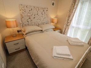 A neatly made bed with a floral headboard, two bedside tables with lamps, and a window with curtains. - Number 15 (Carnforth)