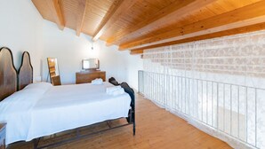 Een slaapkamer met een houten plafond, een bed met witte lakens, een nachtkastje, een dressoir en een raam met uitzicht op een stenen muur.
