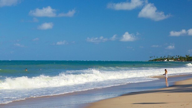 A beach with clear blue water, a person standing on the sand, and a distant shoreline with buildings. - House Maraca (Ipojuca)