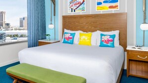 A hotel room with a large bed, a green bench, a nightstand, a lamp, and a view of the cityscape. - Margarittaville Las Vegas  (Las Vegas)
