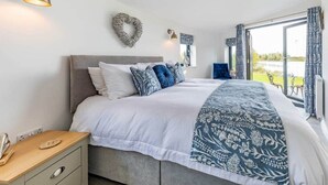 A bedroom with a large bed, a bedside table, a wall-mounted light, a heart-shaped decoration, and a view of the outdoors. - Cosy Luxury Lakeside Lodge, surrounded by amazing views and countryside. (Newark)