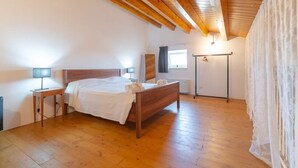 Ein Schlafzimmer mit Holzboden, einem Bett mit weißen Textilien, einem Nachttisch, einem Fenster mit Vorhängen und einem Heizkörper.