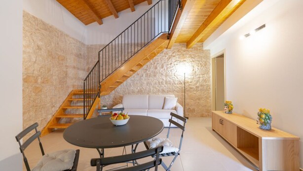 Ein Essbereich mit einem runden Tisch, Stühlen und einer Obstschale. Im Hintergrund ist eine Treppe mit Holzhäuschen und eine weiße Wand zu sehen.