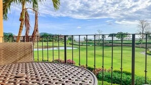 Ein runder Metalltisch mit gewebtem Muster, eine Palme und im Hintergrund ein gepflegter Golfplatz hinter einem schwarzen Metallzaun.