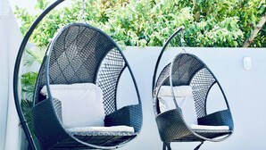 Two black wicker chairs with white cushions, positioned outdoors against a white wall with a green hedge in the background.