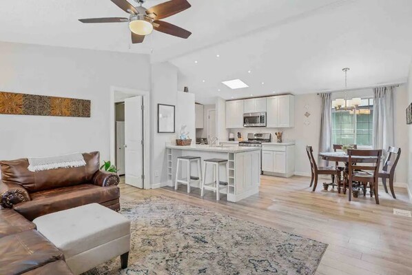 A modern living room with a brown leather sofa, a white ottoman, and a patterned rug. In the background, there is a kitchen with white cabinets, a dining area with a wooden table and chairs, and a ceiling fan.