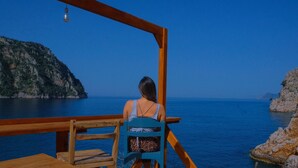 A woman sitting on a wooden deck chair overlooking a body of water with cliffs on one side and a wooden railing. - Kelebekler Vadisi Butterfly Valley (Fethiye)