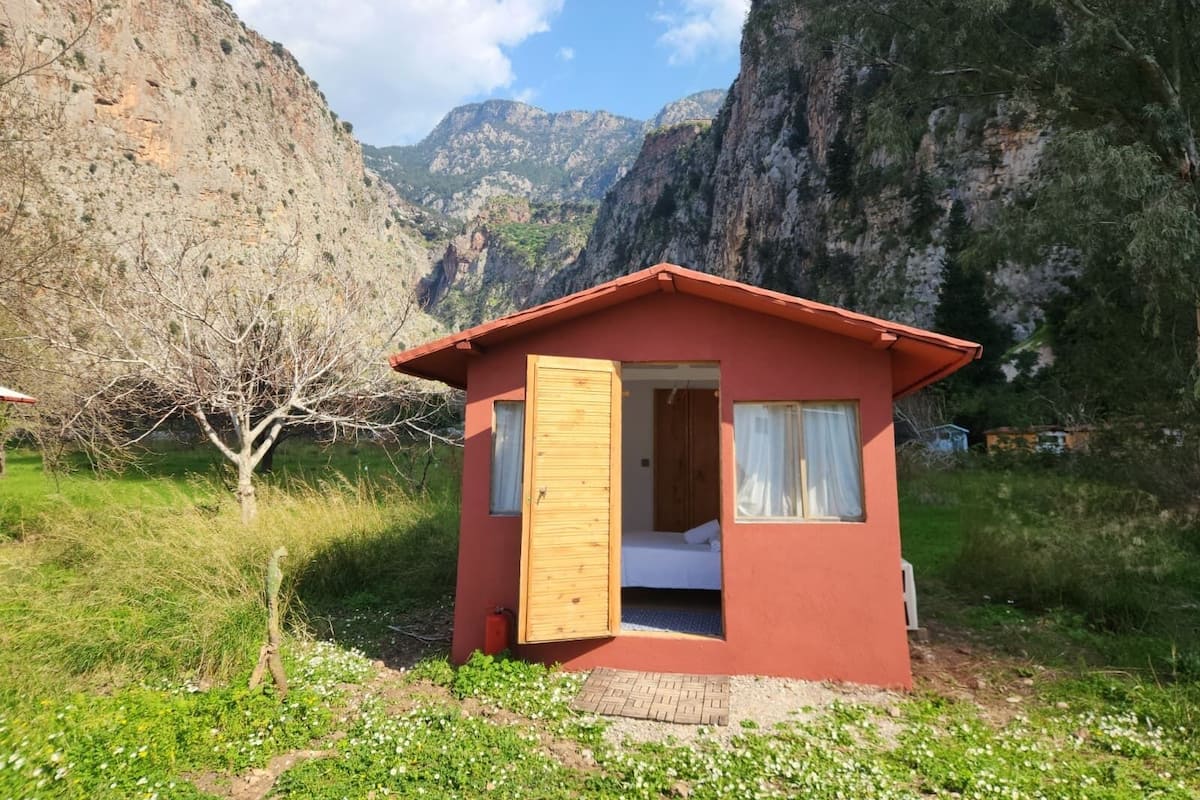 Una cabaña roja pequeña con puerta y ventanas de madera, rodeada de montañas y vegetación.
