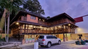 Ein zweistöckiges Holzhaus mit Balkon, einem geparkten Auto davor und einem roten Markisenvorbau auf der rechten Seite.