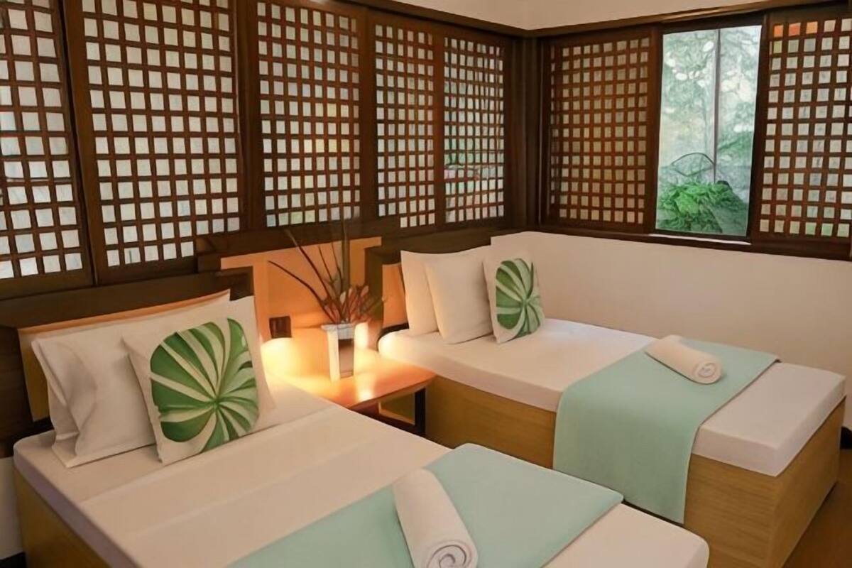 A room with two beds, a small table, and a window with a view of greenery.