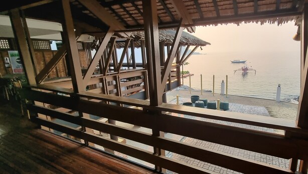 Eine Holzterrasse mit Blick auf das Meer, ein Gebäude mit Strohdach und ein Boot im Wasser.