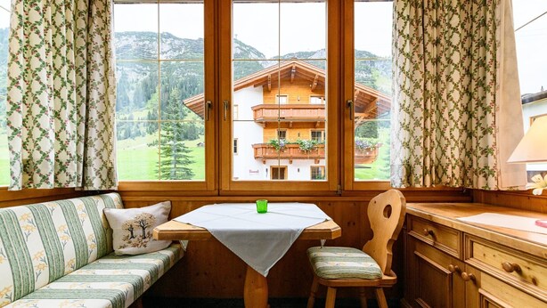 A cozy room with a wooden table and bench, a window with a view of a mountain house, and curtains with a floral pattern. - Montfort (Lech am Arlberg)