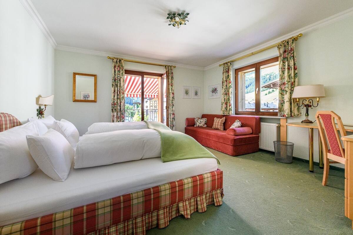 A room with a bed, a red sofa, a small table, and a chair. There are curtains on the windows, and a picture frame on the wall.