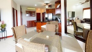 A modern kitchen with wooden cabinets and a central island, a dining area with wicker chairs and a glass table, and a living room with a sofa and a TV.
