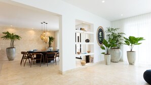 A modern living room with a dining area, featuring a marble floor, large potted plants, and a contemporary light fixture.