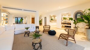 A spacious living area with a white sofa, a wicker chair, and a round black ottoman. There are potted plants and a small wooden table with decorative items. - Casa Delfín by Luxe Haus™  – Butlered Villa, Staff, Pool, Golf Carts (La Romana)