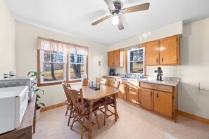 Una cocina con armarios de madera, una mesa de comedor con sillas, un ventilador de techo y una ventana con vistas a árboles.