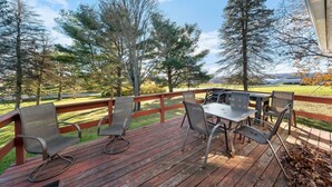 A wooden deck with metal chairs and a table, overlooking a grassy area with trees and a distant view. - Countryside- 3 Bedroom Home (Mantua)