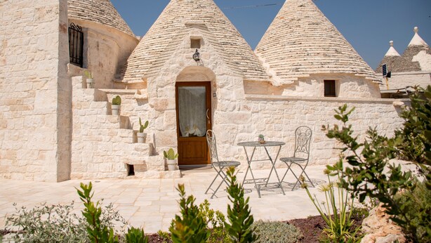 A stone building with conical roofs, a wooden door, and a patio with metal chairs. - Trullo manivida Mulberry (Locorotondo)